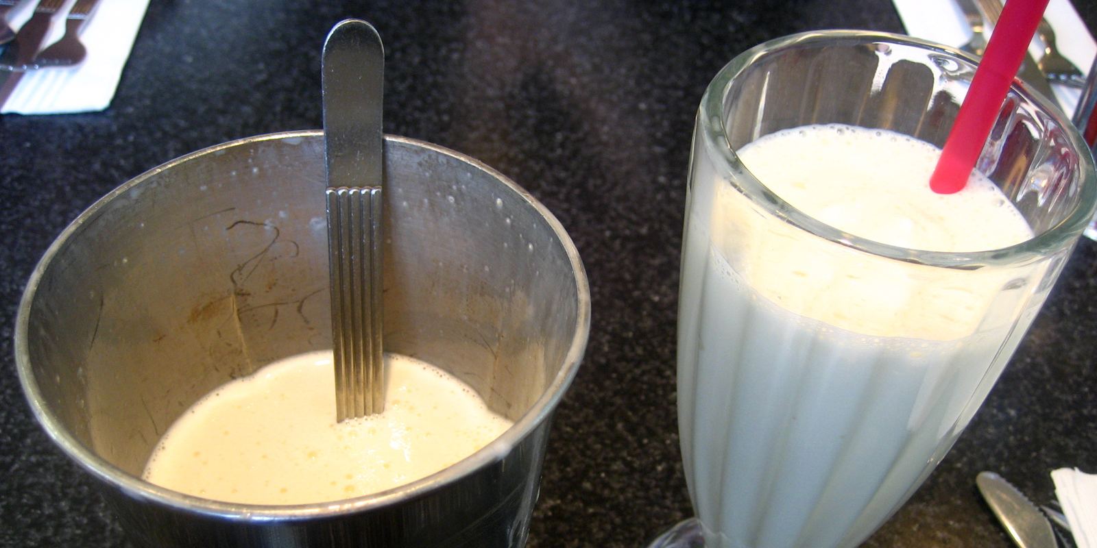 A tall milkshake glass with a metal mixing cup beside it